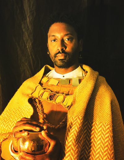 Brathwaite dressed in yellow and gold fabrics, bathed in yellow light, clasping small vase with shiny surface