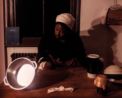 Brathwaite in dark garment with white cloth head covering, bending over kitchen work surface with crockery and a pan; Bach score in the background