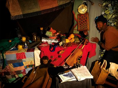 Fabric draped tables covered with dishes, food, and drink; musical instruments and baggage in foreground; at right Brathwaite looking on with expression of glee