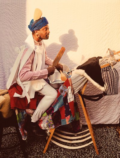 Brathwaite sits on a fabric-draped wooden clothes horse; he has two pans resting in front of him like drums and he holds wooden kitchen utensils as drumsticks