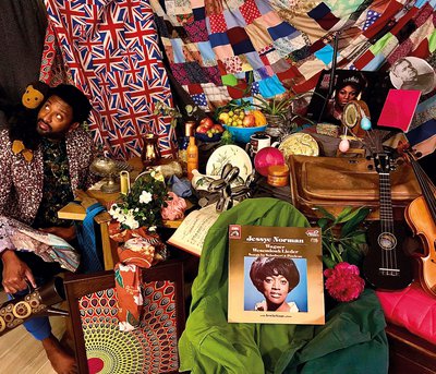 Fabric hangings are backdrop to table covered with numerous different objects; at left Brathwaite sits looking out of frame top left, with stuffed toy monkey on his right shoulder