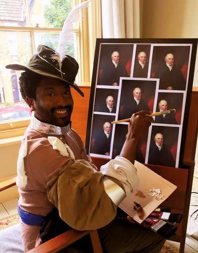 Grinning Brathwaite looks directly out of frame at viewer; he holds paintbrush in right hand and sits in front of easel covered with many copies of a portrait of Victorian era gentleman